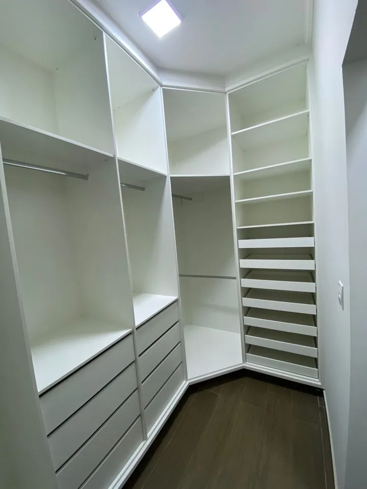 Closet planejado em MDF branco em formato de canto, com cabideiros, gaveteiro central e sapateira em prateleiras abertas