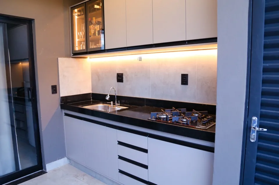 Espaço gourmet em MDF cinza claro com cristaleira de vidro iluminada, revestimento em cimento queimado e bancada de granito preto com cooktop