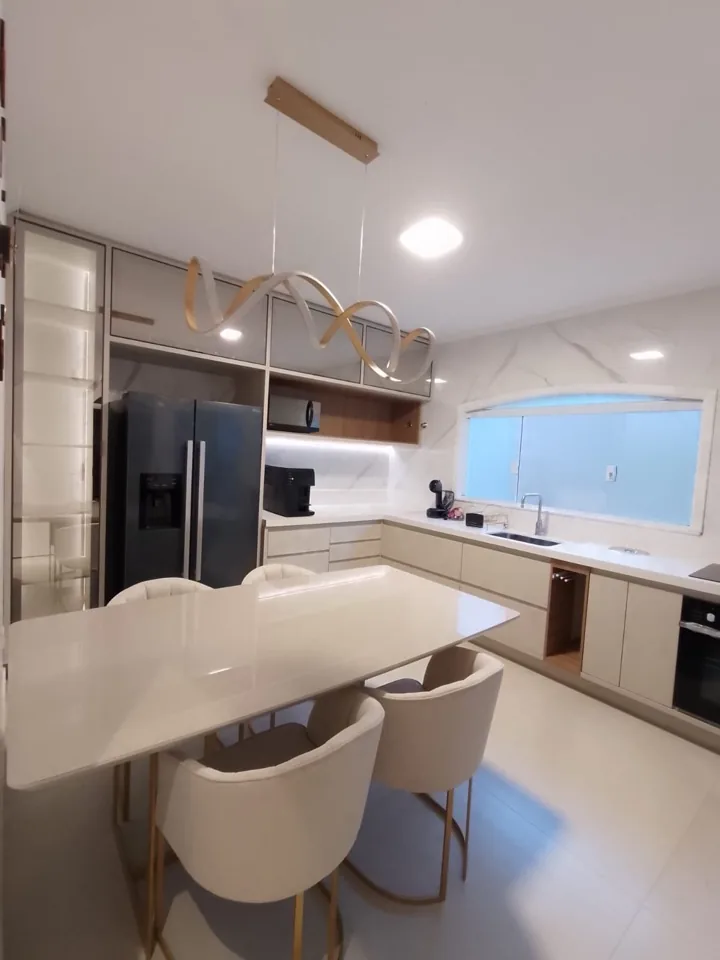 Cozinha em L off-white com mesa de jantar integrada, pendente escultural dourado, geladeira side by side preta e bancada de mármore branco