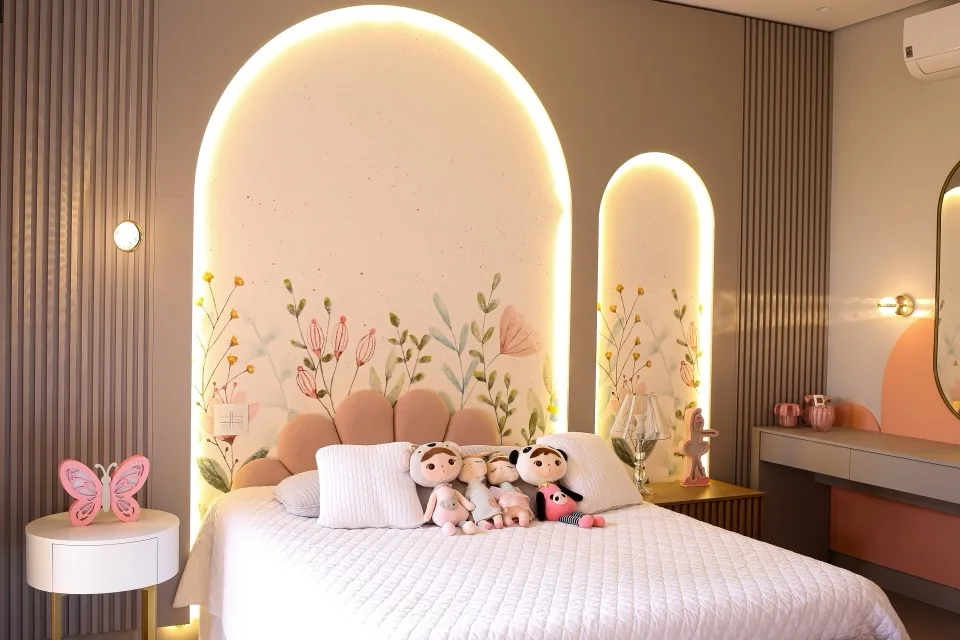 Quarto infantil feminino com cabeceira estofada em pétalas rosa sobre nicho arqueado iluminado com mural floral, painel ripado bege e penteadeira planejada