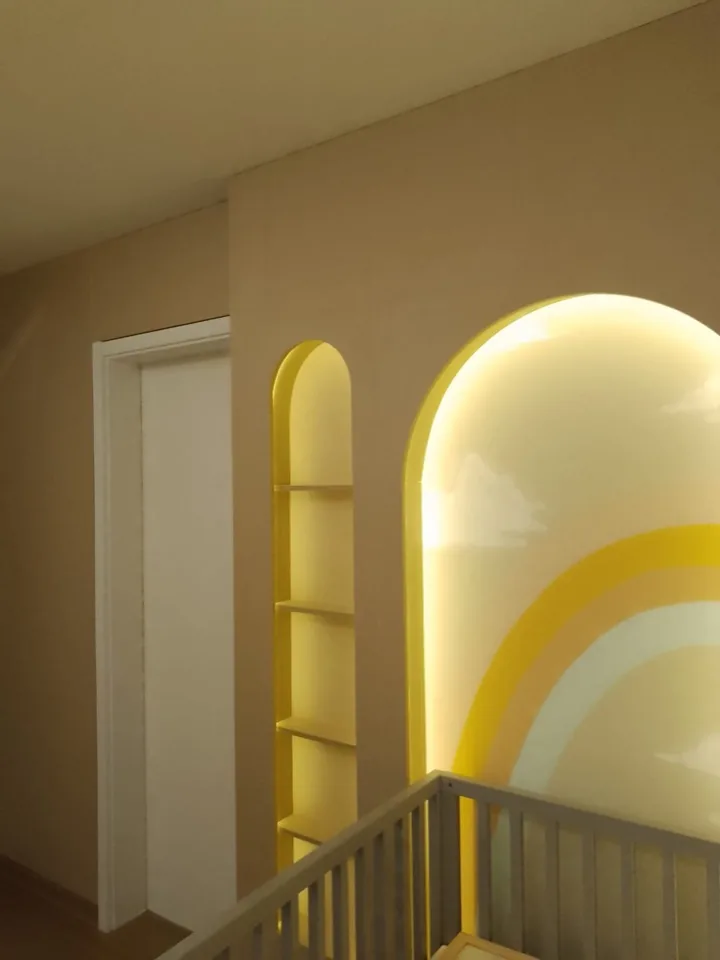 Nicho arqueado planejado em laca amarela com prateleiras e iluminação embutida, ao lado de arco iluminado com mural de arco-íris em quarto de bebê