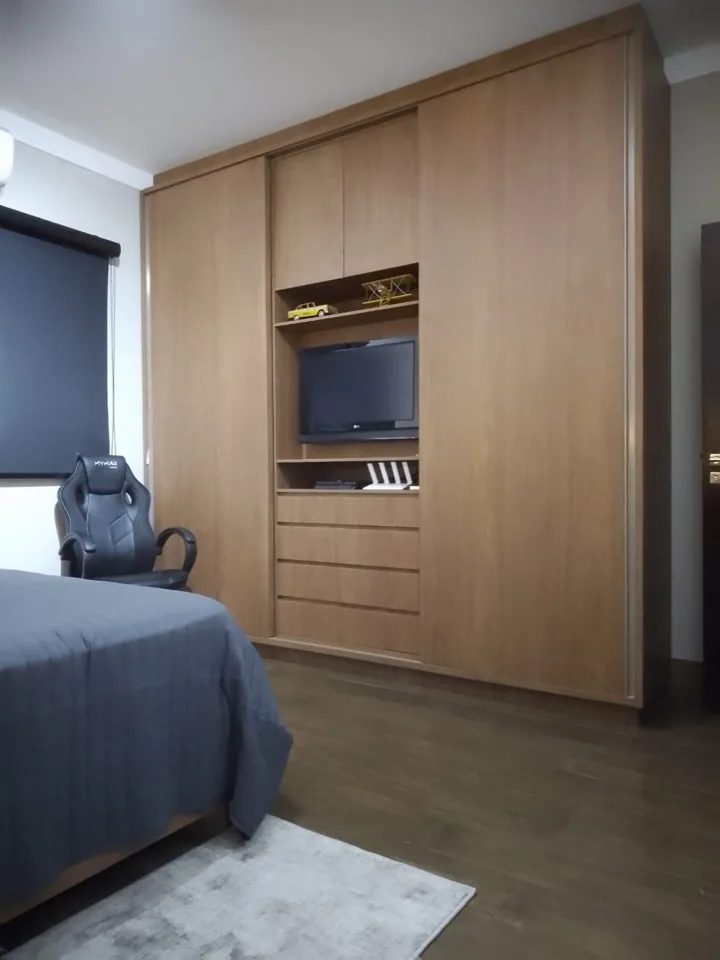 Guarda-roupa planejado de correr em madeira mel ocupando a parede inteira, com nicho central para TV, prateleiras e gaveteiro, em quarto juvenil de piso amadeirado escuro